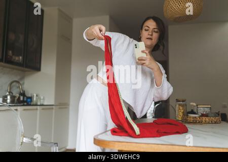 Eine Frau nimmt ein Foto ihres frisch gestrickten roten Pullovers mit einem Maßband in der Nähe auf und kombiniert ihre Liebe zum Stricken und Technologie in einem gemütlichen Zuhause Stockfoto