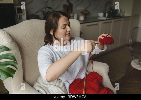 Eine Frau strickt einen leuchtend roten Pullover, während sie bequem in einem gemütlichen Zimmer sitzt. Die Szene fängt die Essenz von Entspannung und Kreativität ein Stockfoto