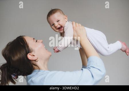 Eine fröhliche Mutter hebt ihr lächelndes Baby in die Luft und schafft einen herzerwärmenden Moment voller Liebe und Zuneigung. Das Baby, in weicher Kleidung gekleidet, genießt es Stockfoto