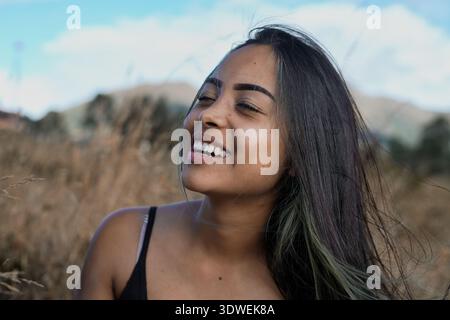 Glückliche junge lateinfrau mit langen dunklen Haaren und Highlights lächelt mit geschlossenen Augen und genießt die frische Luft in einem friedlichen Feld mit Bergen Stockfoto