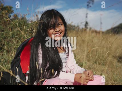Glückliches indigenes Mädchen mit langen dunklen Haaren und rosafarbenen Kleidern, das mit ihrem Rucksack auf einem Feld aus hohem Gras sitzt, das sonnige Wetter genießt und lächelt Stockfoto