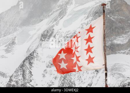 Vor dem Hintergrund der verschneiten Berge in den Schweizer Alpen bei Zermatt weht eine lebendige Walliser Kantonsflagge. Die roten und weißen Sterne stehen im Kontrast Stockfoto