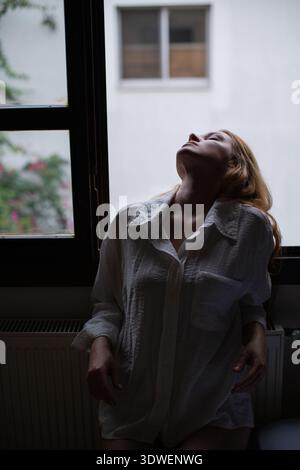 Ein kontemplatives Porträt einer Frau, die in einem dunklen Raum vor einem Fenster steht. Die sanfte Beleuchtung und der Kontrast erzeugen eine Stimmung der Introspektion und des Geheimnisses Stockfoto
