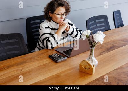 Frau mittleren Alters, die an einem Holzschreibtisch in einem modernen Büro arbeitet und sich auf ihren Laptop konzentriert. Bürobedarf und Blumen schmücken den Arbeitsplatz, kreativ Stockfoto