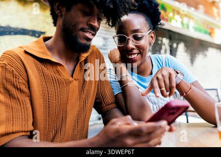 Ein fröhliches Paar, das einen Moment zusammen genießt und auf einen Smartphone-Bildschirm blickt. Sie sitzen in einem Café im Freien, teilen ein Lächeln und diskutieren, was es ist Stockfoto