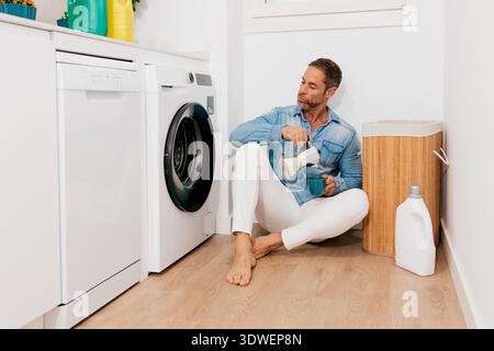 Ein Mann in einem lichtdurchfluteten Waschraum genießt eine Tasse Kaffee, während er auf dem Boden sitzt. Umgeben von Wäsche-Essentials sorgt er für Entspannung Stockfoto