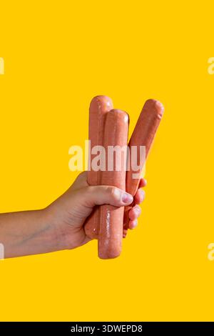 Eine Hand hält drei Würstchen vor einem leuchtend gelben Hintergrund. Die kontrastierenden Farben betonen das Motiv und bieten ein verspieltes und appetitliches Bild Stockfoto