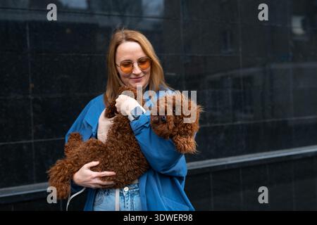 Eine lächelnde Frau in einer blauen Jacke hält ihren flauschigen braunen Hund in der Hand und genießt eine ungezwungene Outdoor-Umgebung. Ihre liebevolle Geste hebt das besondere b hervor Stockfoto