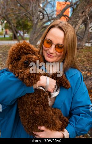 Eine fröhliche Frau mit orangefarbener Sonnenbrille hält ihren flauschigen braunen Hund im Park. Umgeben von Herbstlaub teilt sie einen freudigen Moment mit ihrem Be Stockfoto
