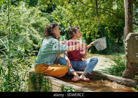 Zwei Schwestern sitzen an einem ruhigen Teich, umgeben von üppigem Grün, und genießen die Gesellschaft des anderen. Einer weist auf etwas Aufregendes in einer Silberbucke hin Stockfoto