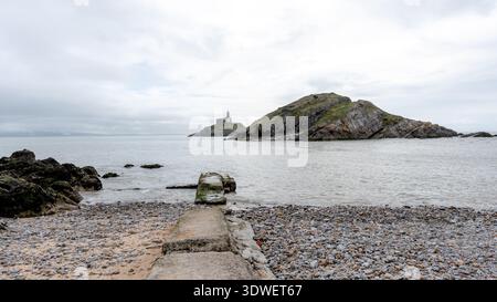 Malerische Aussicht auf den Mumbles Lighthouse vom Kiesstrand in Bracelet Bay, erfasst an einem bewölkten Tag mit üppigem Grün auf der felsigen Insel, die einen schafft Stockfoto
