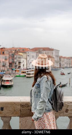 Rückansicht einer unerkennbaren Frau, die auf der Rialto-Brücke steht, mit Blick auf den Canal Grande in Venedig, Italien, mit Hut und Jeansjacke, die sie festnimmt Stockfoto