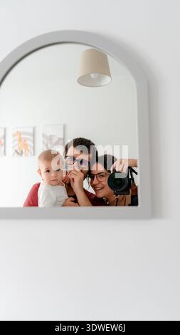 Eine fröhliche Familie fängt einen kostbaren Moment in einem Spiegel ein. Das Baby wird von den Eltern gehalten, während die Mutter geschickt die Erinnerung mit einfängt Stockfoto