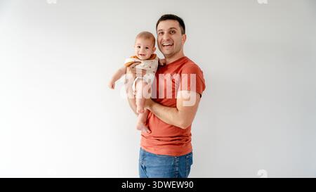 Ein fröhlicher Vater hält sein lächelndes Baby in warmer Umarmung vor einem einfachen Hintergrund. Die Szene fängt einen zärtlichen Familienmoment voller Glück und ein Stockfoto