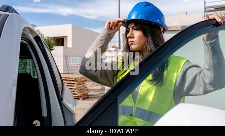 Ein technischer Techniker mit blauem Schutzhelm und reflektierender Weste überprüft den Fortschritt auf einer Baustelle. Sie steht neben einem Fahrzeug und bewertet das Stockfoto