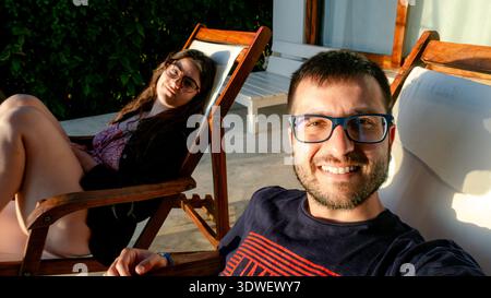 Ein Paar genießt einen ruhigen Sonnenaufgang auf einer privaten Hotelterrasse in Sansibar, Tansania, in hölzernen Liegestühlen. Sie sind von üppigem Grün umgeben Stockfoto