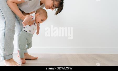 Eine Mutter stützt ihr fünfmonatiges Baby sanft, während er lernt, auf einem Holzboden zu stehen. Die Szene fängt einen zarten Moment des Wachstums und der Bindung ein Stockfoto