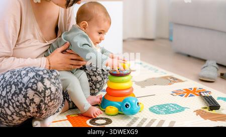 Das Baby sitzt auf einer gemusterten Matte und spielt mit einem bunten, stapelbaren Spielzeug, das von einem Erwachsenen überwacht wird. Die Umgebung ist entspannt, was die frühkindliche P Stockfoto
