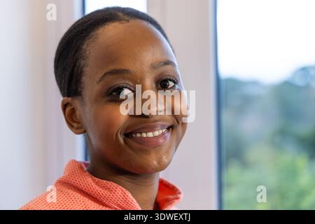 Afroamerikanische Teenager-Frau lächelt und sieht in die Kamera am Fenster mit korallenrosafarbenem Reißverschluss Stockfoto