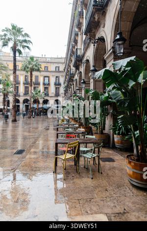 Leerer Sitzbereich im Freien eines Cafés in Barcelona, Spanien, mit nassem Bürgersteig, der den bewölkten Himmel und die umliegende Architektur reflektiert Stockfoto