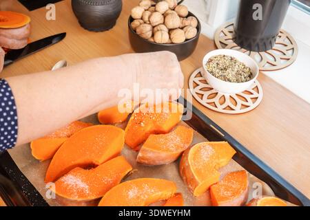Eine Frau würzt Kürbis in Scheiben mit Gewürzen in der Küche und bereitet ihn zum Backen vor. Eine gemütliche, warme Atmosphäre mit frischen Zutaten auf einer Holztheke Stockfoto