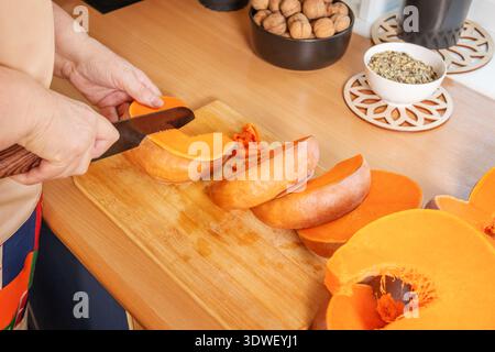 Eine Frau schneidet einen Kürbis auf einem hölzernen Schneidebrett in ihrer Küche und bereitet ihn zum Backen vor. Nüsse und Körner werden in Schüsseln auf den Zähler gelegt Stockfoto