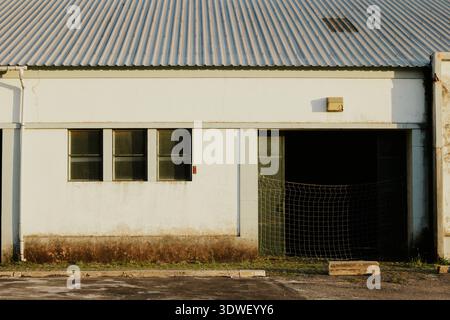 Ein altes Lagerhaus mit einer verwitterten Fassade und einem Wellblechdach unter warmem Sonnenlicht. Der Eingang, teilweise mit einem Netz bedeckt, fügt eine Industrie hinzu Stockfoto