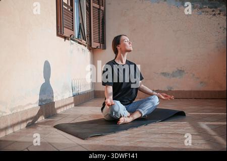 Eine Person sitzt im Kreuz auf einer Yogamatte und meditiert unter der Morgensonne auf einem Balkon. Diese Szene fängt das Wesen eines ruhigen und achtsamen morgens ein Stockfoto