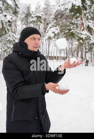Ein Mann in schwarzem Wintermantel und Hut jongliert Schneebälle in einem verschneiten Wald. Die ruhige Winterlandschaft ist voller schneebedeckter Bäume und einer Stockfoto