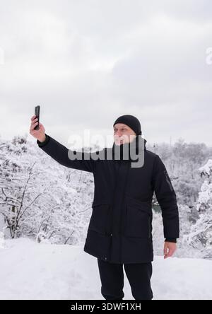 Ein Mann in schwarzer Winterjacke und Hut macht ein Selfie vor dem Hintergrund einer schneebedeckten Landschaft. Die Szene fängt die heitere Schönheit des Winters ein Stockfoto
