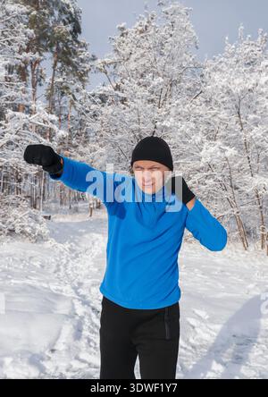 Ein Mann in blauer Jacke und schwarzen Handschuhen trainiert draußen in einem verschneiten Wald. Er konzentriert sich auf seine Trainingsroutine und nimmt den frischen Winterenvir auf Stockfoto