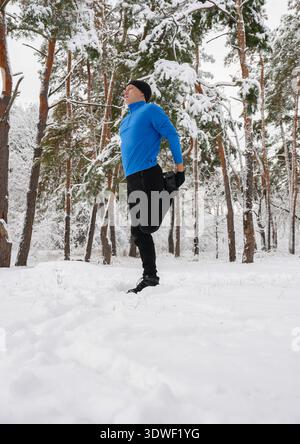Ein Mann in sportlicher Kleidung dehnt sein Bein in einem verschneiten Wald und bereitet sich auf ein Wintertraining vor. Die Bäume sind mit Schnee bedeckt und schaffen eine ruhige Kulisse Stockfoto