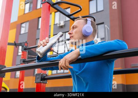 Ein Mann, der Kopfhörer und eine blaue Jacke trägt, macht beim Sport im Freien eine Trinkpause. Er lehnt sich an dunklen Bars vor einem pulsierenden Gebäude und genießt es Stockfoto