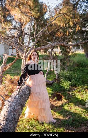 Eine junge schwangere Frau steht friedlich in einer natürlichen Umgebung und lehnt sich an einen Baum. Ihr sanfter Ausdruck spiegelt die Schönheit und Ruhe von Mo wider Stockfoto