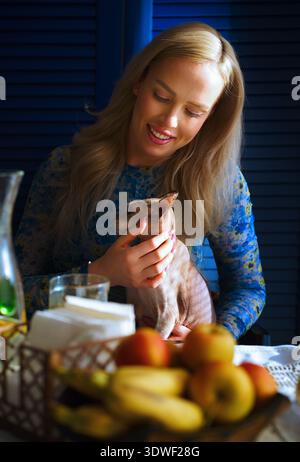 Eine blonde Frau kuschelt ihre Sphynx-Katze sanft in einer gemütlichen Umgebung, umgeben von einem warmen Ambiente. Ihre Bindung spiegelt Liebe und Ruhe in Th wider Stockfoto