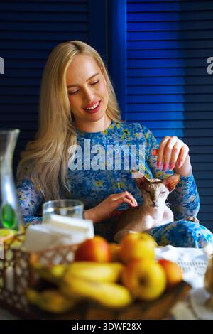 Eine blonde Frau sitzt zu Hause mit ihrer Sphynx-Katze auf dem Schoß. Sie lächelt herzlich und genießt einen friedlichen Moment in einer gemütlichen häuslichen Umgebung voller n Stockfoto