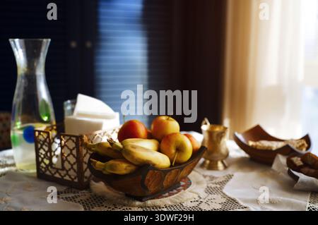 Ein rustikaler Tisch ist mit einer Holzschale mit frischen Bananen und Äpfeln geschmückt, die in einem sanft beleuchteten Raum an eine heimelige, gesunde Lifestyle-Szene erinnert. Stockfoto