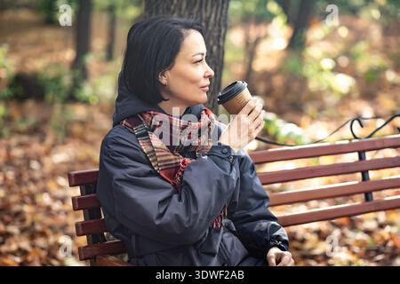 Eine Frau sitzt auf einer Parkbank und genießt an einem Herbsttag eine Tasse Kaffee. Sie ist in einen kuscheligen Schal gewickelt, umgeben von den warmen Tönen gefallener Blätter, Stockfoto