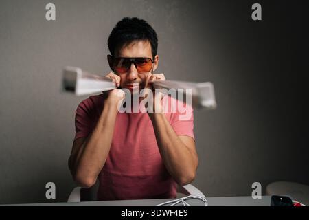 Porträt eines stilvollen jungen erwachsenen Mannes, der ein rosafarbenes Hemd und eine getönte Sonnenbrille trägt und LED-Lichtbalken über den Schultern hält, Gesicht und Licht beleuchtet Stockfoto