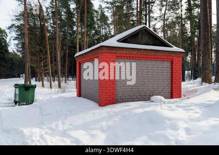 Funktioneller, roter und grauer Ziegelschuppen und ein grüner Abfalleimer, der in tiefem Schnee in der Nähe einer Forststraße steht. Facility Management und Versorgungsleistungen im Winter. Stockfoto