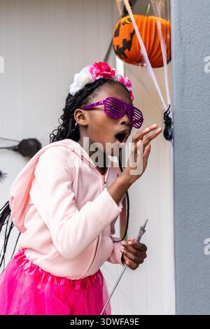 Afroamerikanisches Kind mit rosa Hoodie und Tutu, das auf die Plastikspinne auf der Veranda streckt Stockfoto