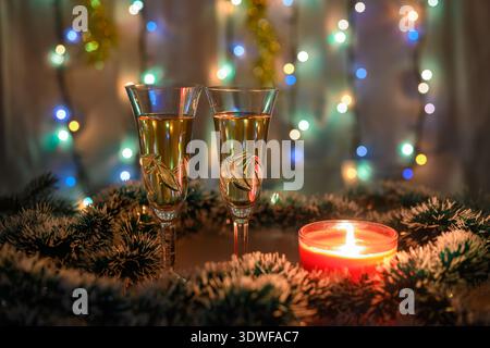 Zwei Gläser Sekt umgeben von grünem und weißem Lametta mit brennender Kerze und einer leuchtenden Girlande im Hintergrund des Neujahrs. Flaches DE Stockfoto