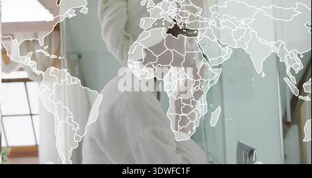 Hautpflegefrau mit Bademantel und Handtuch-Turban am Waschtisch im Bad, Spiegel, Weltkarte-Overlay Stockfoto