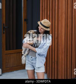 Lächelnde junge Frau in Strohhut, die einen kleinen süßen Hund hält, der auf der Veranda eines modernen Hauses mit einer Holzfassade steht, die glücklichen Lebensstil repräsentiert, Immobilien Stockfoto