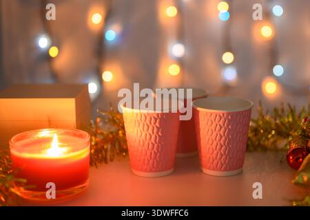 Drei pinkfarbene Tassen auf weihnachtlichem Hintergrund mit brennenden Kerzen und einer leuchtenden Girlande in Orange. Geringe Schärfentiefe. Stockfoto