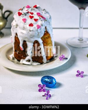 Traditioneller orthodoxer Osterkuchen (Kulich) mit weißem Zuckerguss und Streuseln, serviert mit zwei Gläsern Rotwein auf dunklem Hintergrund. Stockfoto