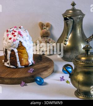 Action-Shot von Milch, die aus einem Milchkännchen in eine Kaffeetasse gegossen wird. Traditioneller Osterkuchen, Fliederblüten und ein Plüschhase im Rock zum Frühstück Stockfoto