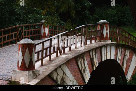Klassische, bogenförmige Ziegelbrücke mit dekorativen Säulen mit Rautenmuster und Holzgeländern. Historischer Kopfsteinpflasterweg über einen Fluss in einem Sommerpark Stockfoto