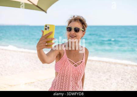 Eine reife Frau genießt einen sonnigen Tag am Strand mit Sonnenbrille und rosa Kleidung. Sie lächelt, während sie ein Selfie macht, den Ozean und einen Onkel einfängt Stockfoto
