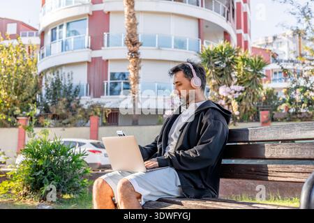 Ein Mann mit Hoodie und Kopfhörern sitzt auf einer Bank und konzentriert sich auf seinen Laptop. Er arbeitet draußen vor modernen Wohngebäuden und genießt es Stockfoto